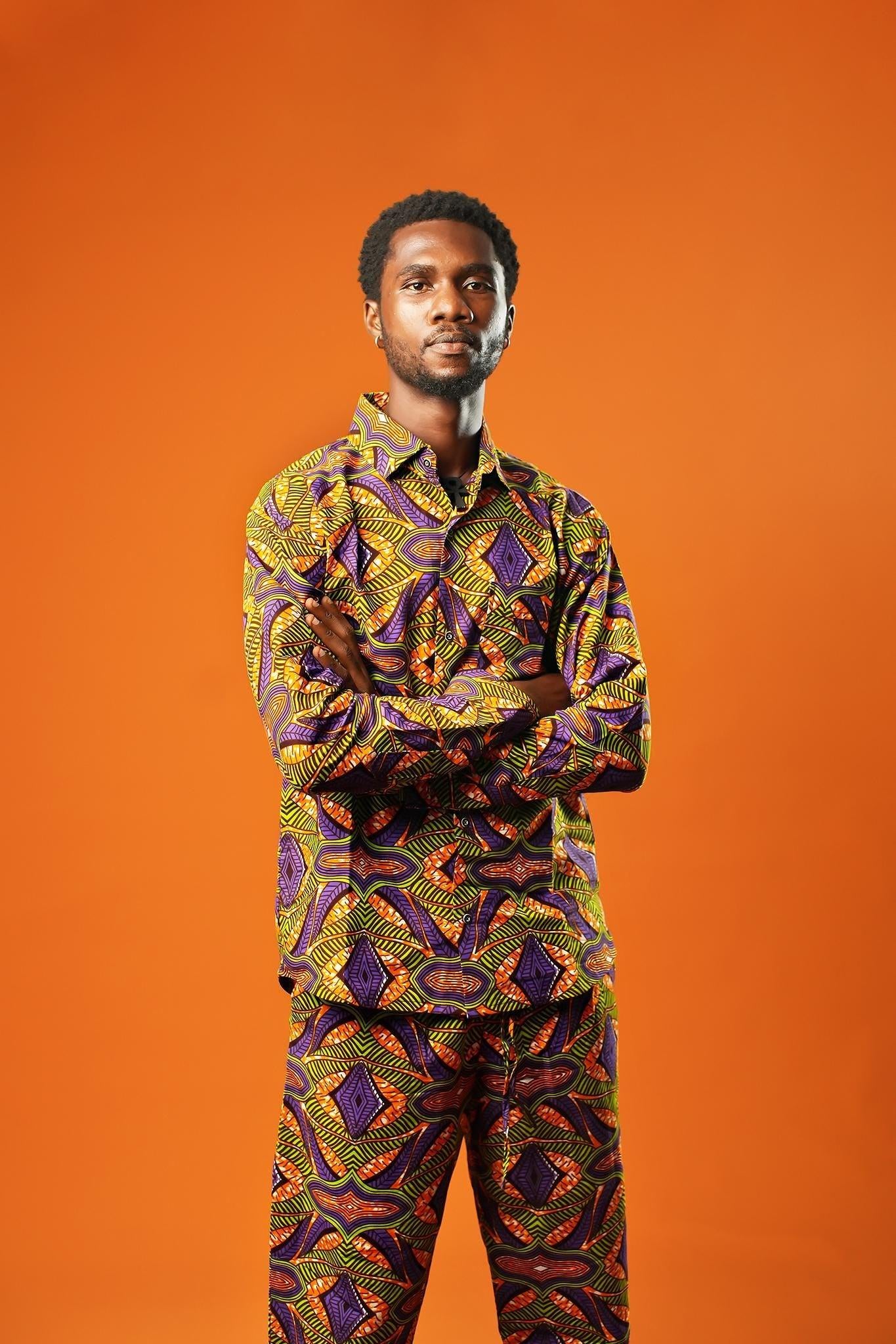 A man stands against an orange background wearing a matching two-piece set made from bold African print fabric. The outfit includes a long-sleeved button-up shirt and straight-leg trousers with a yellow, purple, red, and black geometric pattern. This is a full Ankara outfit for men, suitable for festival fashion or statement African clothing.