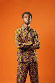 A man stands against an orange background wearing a matching two-piece set made from bold African print fabric. The outfit includes a long-sleeved button-up shirt and straight-leg trousers with a yellow, purple, red, and black geometric pattern. This is a full Ankara outfit for men, suitable for festival fashion or statement African clothing.