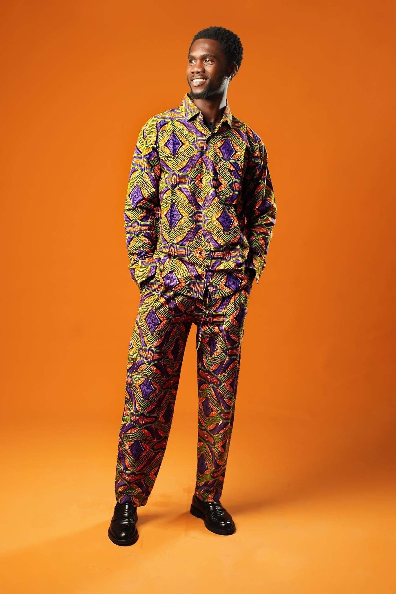 A man stands against an orange background wearing a matching two-piece set made from bold African print fabric. The outfit includes a long-sleeved button-up shirt and straight-leg trousers with a yellow, purple, red, and black geometric pattern. This is a full Ankara outfit for men, suitable for festival fashion or statement African clothing.