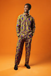 A man stands against an orange background wearing a matching two-piece set made from bold African print fabric. The outfit includes a long-sleeved button-up shirt and straight-leg trousers with a yellow, purple, red, and black geometric pattern. This is a full Ankara outfit for men, suitable for festival fashion or statement African clothing.