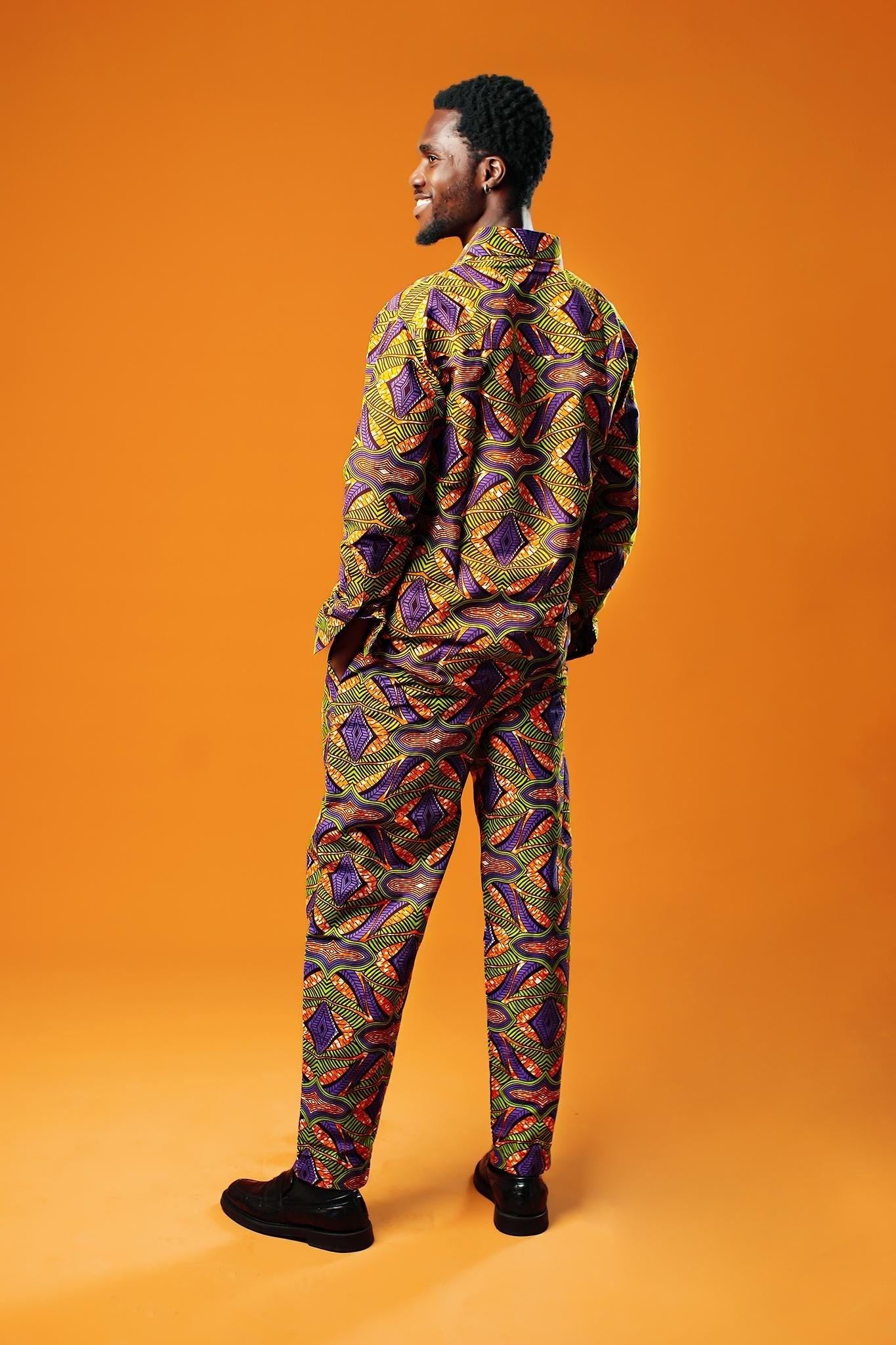 A man stands against an orange background wearing a matching two-piece set made from bold African print fabric. The outfit includes a long-sleeved button-up shirt and straight-leg trousers with a yellow, purple, red, and black geometric pattern. This is a full Ankara outfit for men, suitable for festival fashion or statement African clothing.