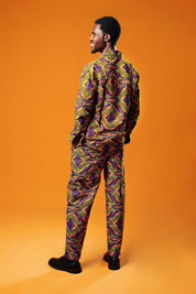 A man stands against an orange background wearing a matching two-piece set made from bold African print fabric. The outfit includes a long-sleeved button-up shirt and straight-leg trousers with a yellow, purple, red, and black geometric pattern. This is a full Ankara outfit for men, suitable for festival fashion or statement African clothing.