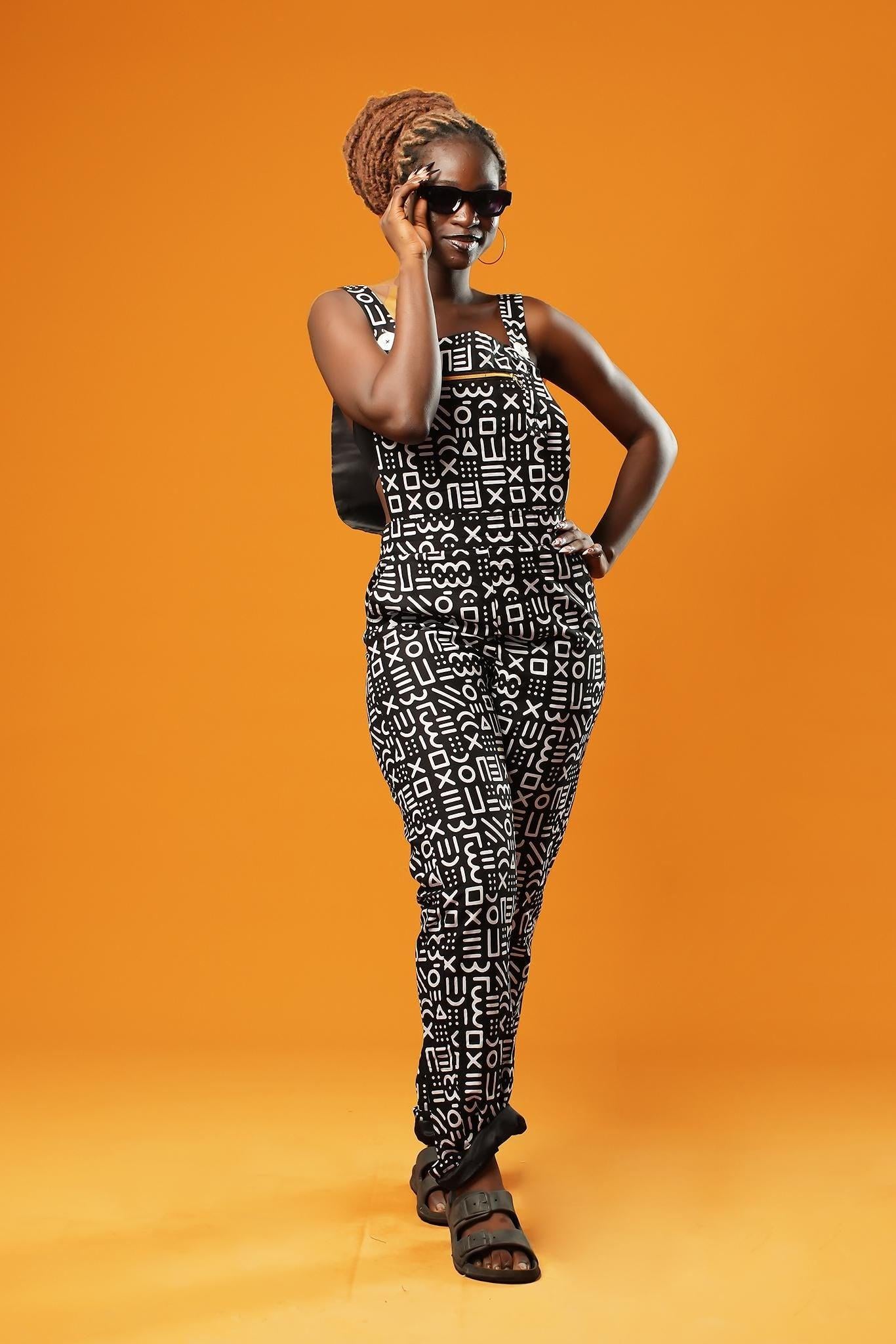 Woman standing against an orange background wearing black and white African print dungarees. The dungarees feature a bold tribal mud cloth pattern and have adjustable straps.