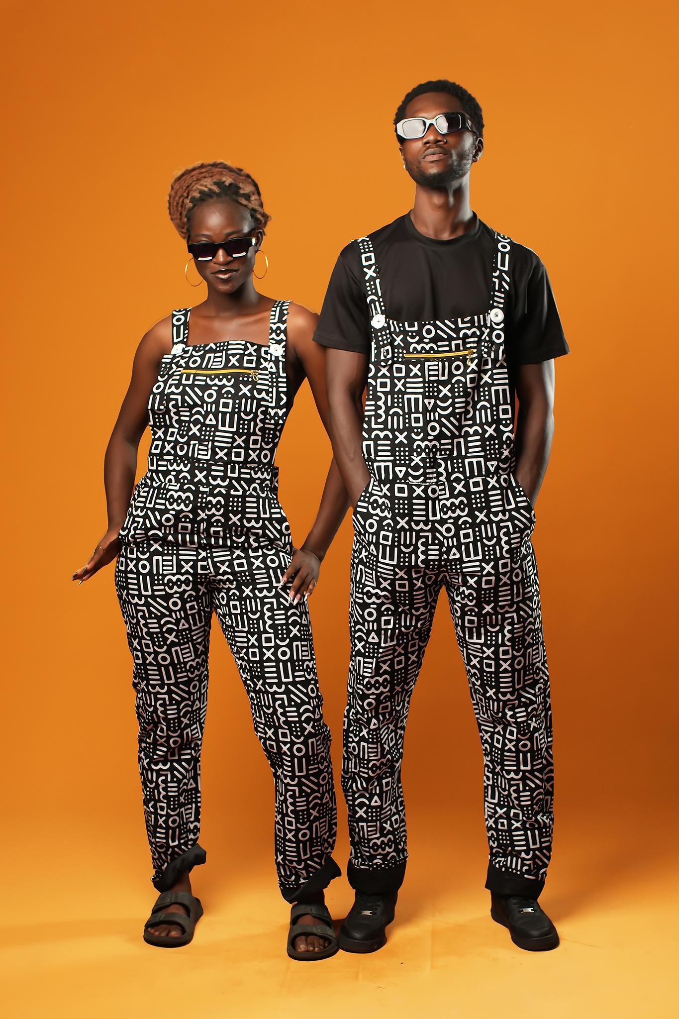 Man and woman standing against an orange background wearing matching black and white African print dungarees. The dungarees feature a bold tribal mud cloth pattern and have adjustable straps.