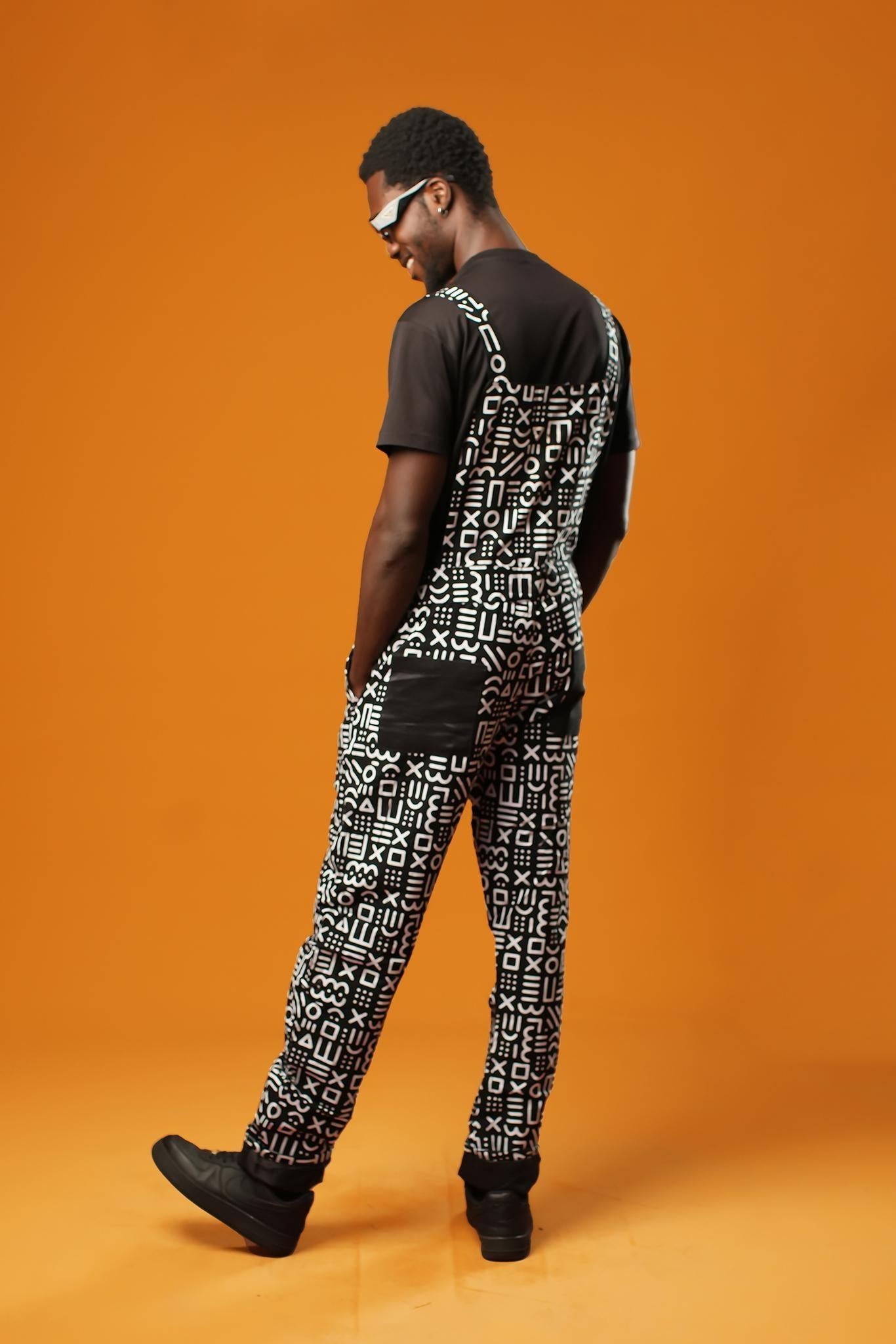 Man standing against an orange background wearing black and white African print dungarees. The dungarees feature a bold tribal mud cloth pattern and have adjustable straps.