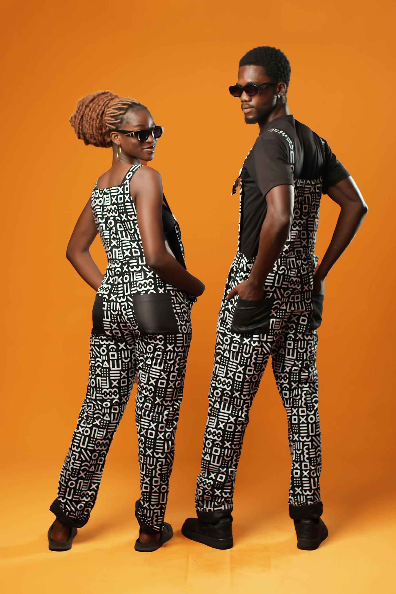 Man and woman standing against an orange background wearing matching black and white African print dungarees. The dungarees feature a bold tribal mud cloth pattern and have adjustable straps.