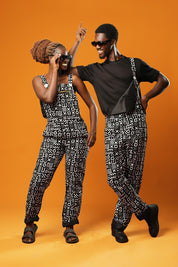 Man and woman standing against an orange background wearing matching black and white African print dungarees. The dungarees feature a bold tribal mud cloth pattern and have adjustable straps.
