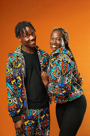 Man and woman wearing matching African print hooded jackets with bold multicoloured geometric patterns. The man is also wearing matching trousers to complete the tracksuit set. Both jackets have front zips and black ribbed cuffs, styled for festival or rave outfits.