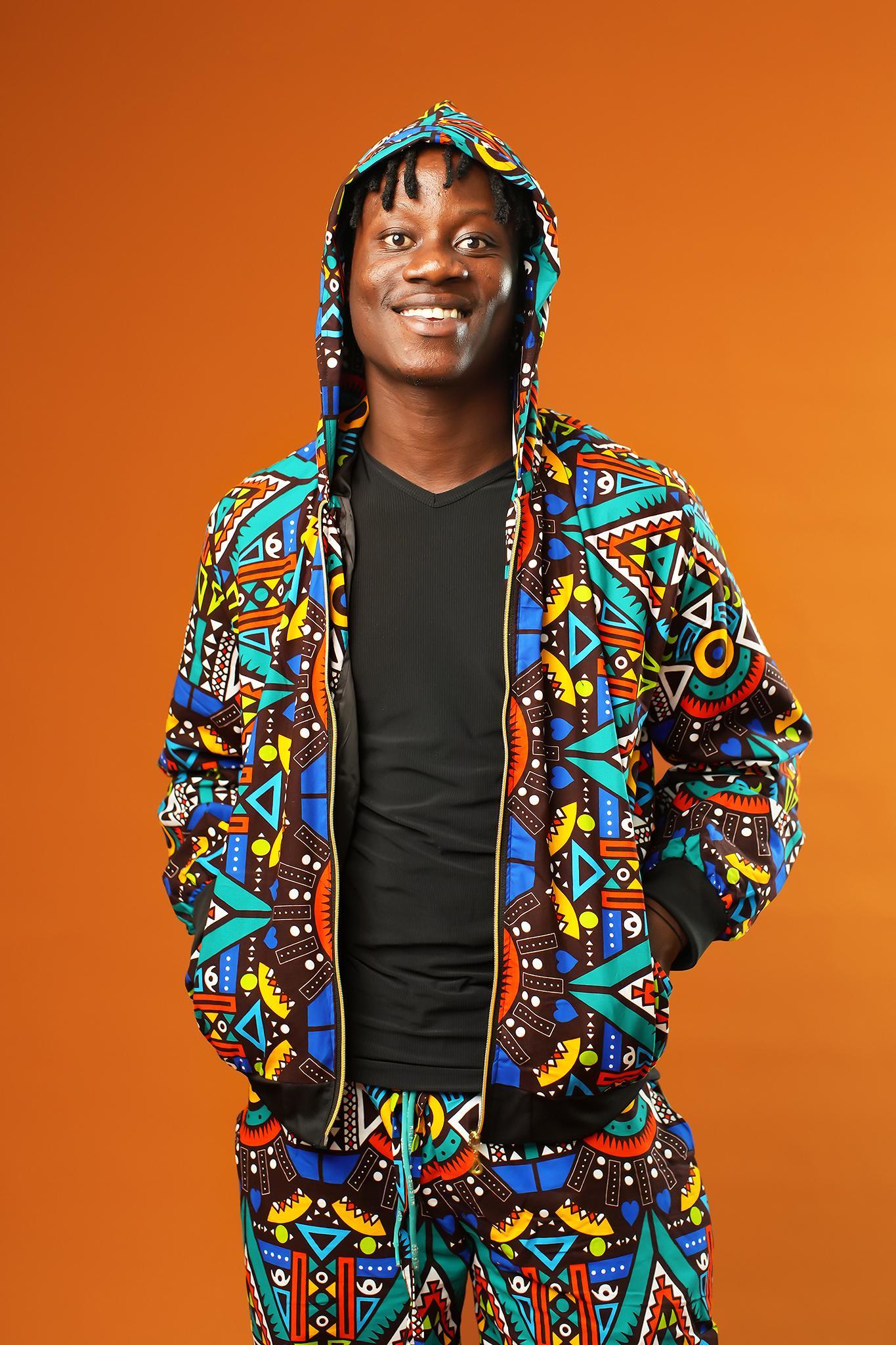 Man wearing African print tracksuit featuring a multicoloured geometric pattern in blue, yellow, red, and black. The outfit includes a zip-up hooded jacket and matching drawstring trousers, styled as a unisex festival or rave set.