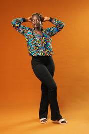 Woman wearing a colourful African print bomber jacket with a bold geometric pattern in blue, red, yellow, and orange. The jacket has a front zip, black ribbed cuffs, and is styled as a rave party hoodie