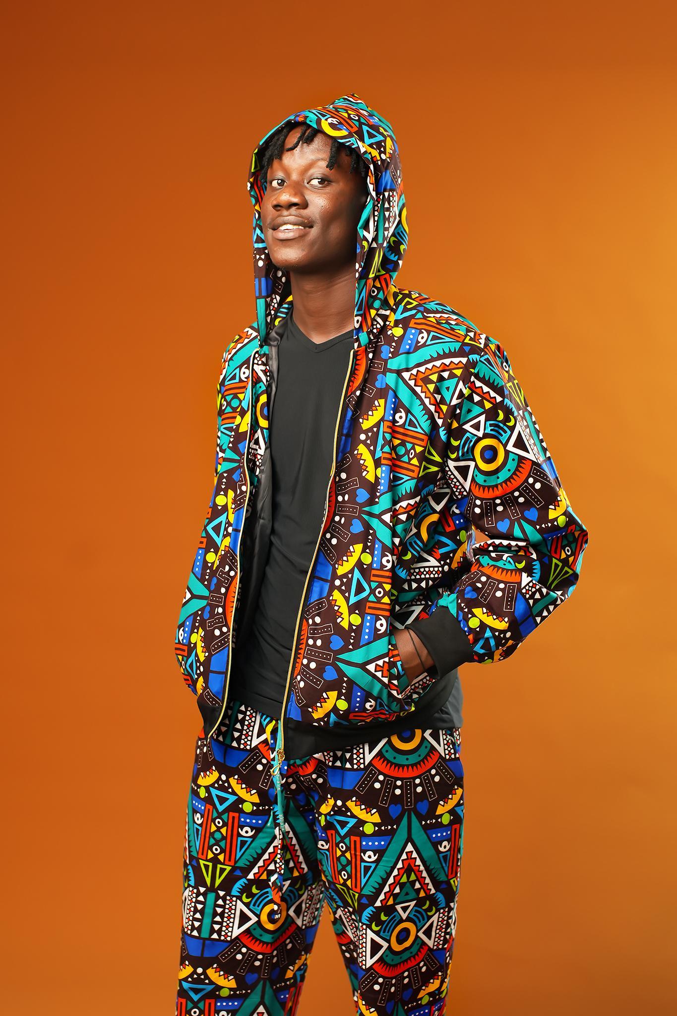 Man wearing African print tracksuit featuring a multicoloured geometric pattern in blue, yellow, red, and black. The outfit includes a zip-up hooded jacket and matching drawstring trousers, styled as a unisex festival or rave set.
