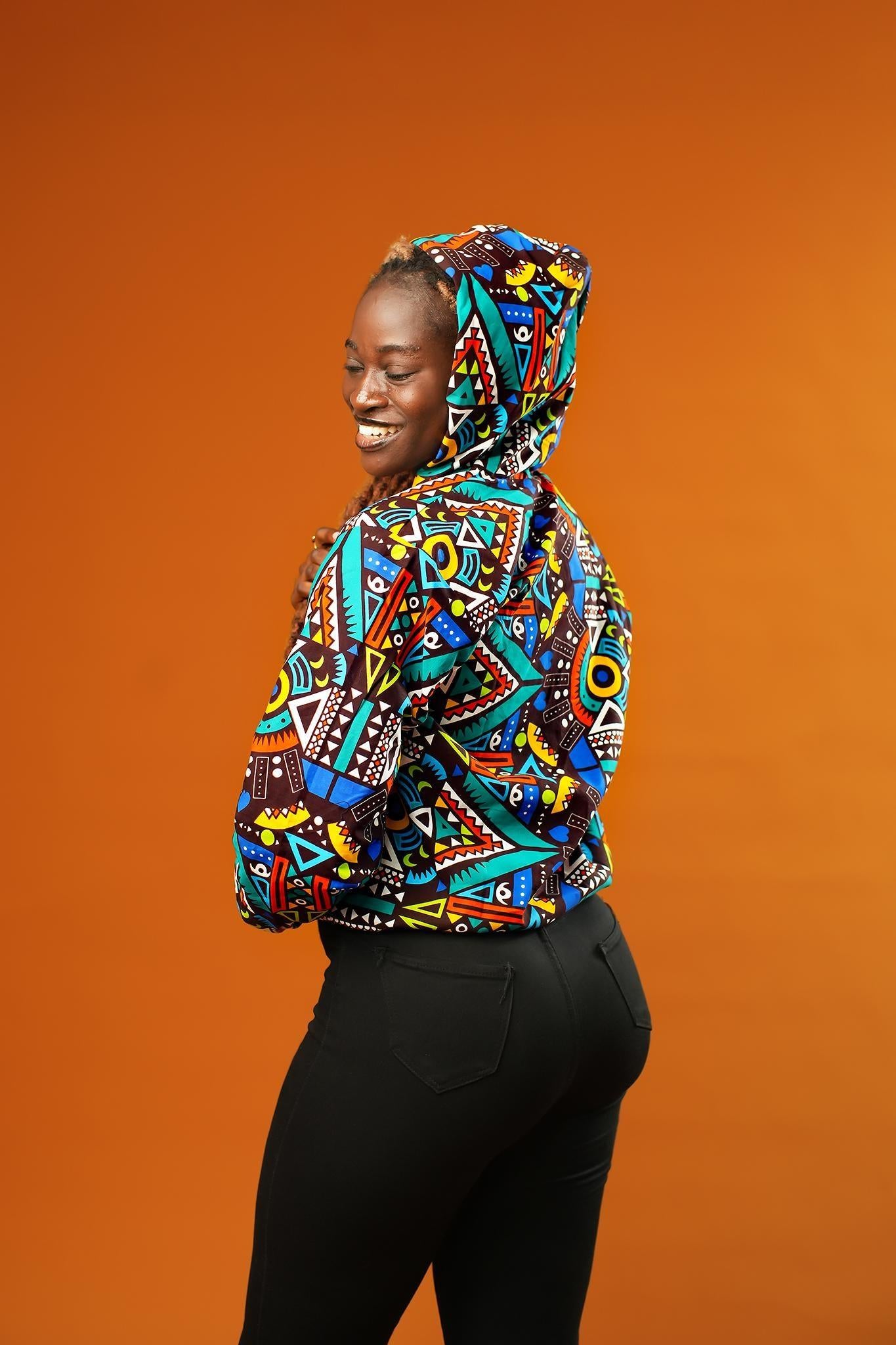 Woman wearing a colourful African print bomber jacket with a bold geometric pattern in blue, red, yellow, and orange. The jacket has a front zip, black ribbed cuffs, and is styled as a rave party hoodie