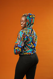 Woman wearing a colourful African print bomber jacket with a bold geometric pattern in blue, red, yellow, and orange. The jacket has a front zip, black ribbed cuffs, and is styled as a rave party hoodie