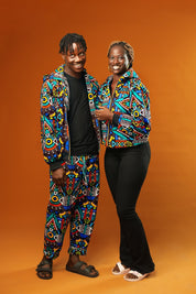 Man and woman wearing matching African print hooded jackets with bold multicoloured geometric patterns. The man is also wearing matching trousers to complete the tracksuit set. Both jackets have front zips and black ribbed cuffs, styled for festival or rave outfits.