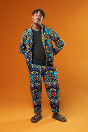 Man wearing African print tracksuit featuring a multicoloured geometric pattern in blue, yellow, red, and black. The outfit includes a zip-up hooded jacket and matching drawstring trousers, styled as a unisex festival or rave set.