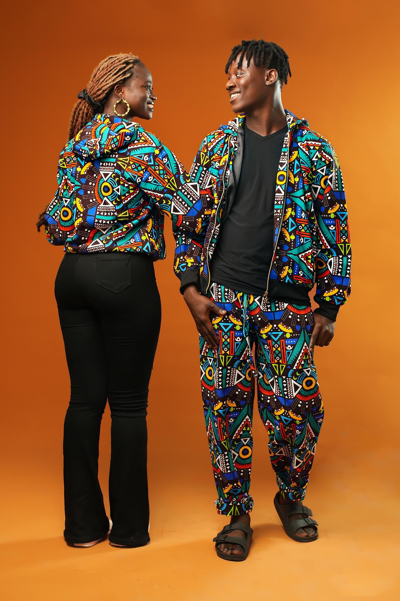 Man and woman wearing matching African print hooded jackets with bold multicoloured geometric patterns. The man is also wearing matching trousers to complete the tracksuit set. Both jackets have front zips and black ribbed cuffs, styled for festival or rave outfits.