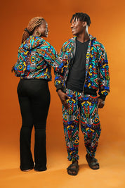 Man and woman wearing matching African print hooded jackets with bold multicoloured geometric patterns. The man is also wearing matching trousers to complete the tracksuit set. Both jackets have front zips and black ribbed cuffs, styled for festival or rave outfits.