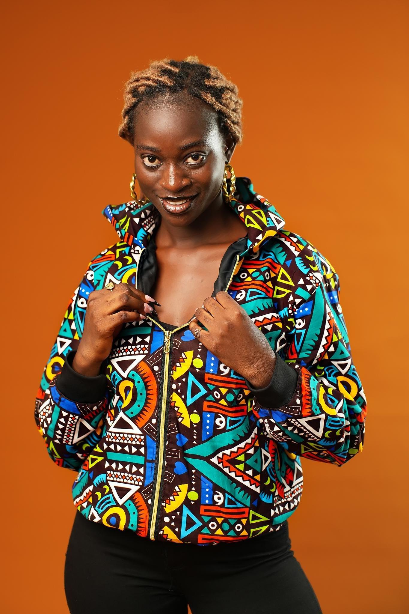 Woman wearing a colourful African print bomber jacket with a bold geometric pattern in blue, red, yellow, and orange. The jacket has a front zip, black ribbed cuffs, and is styled as a rave party hoodie.