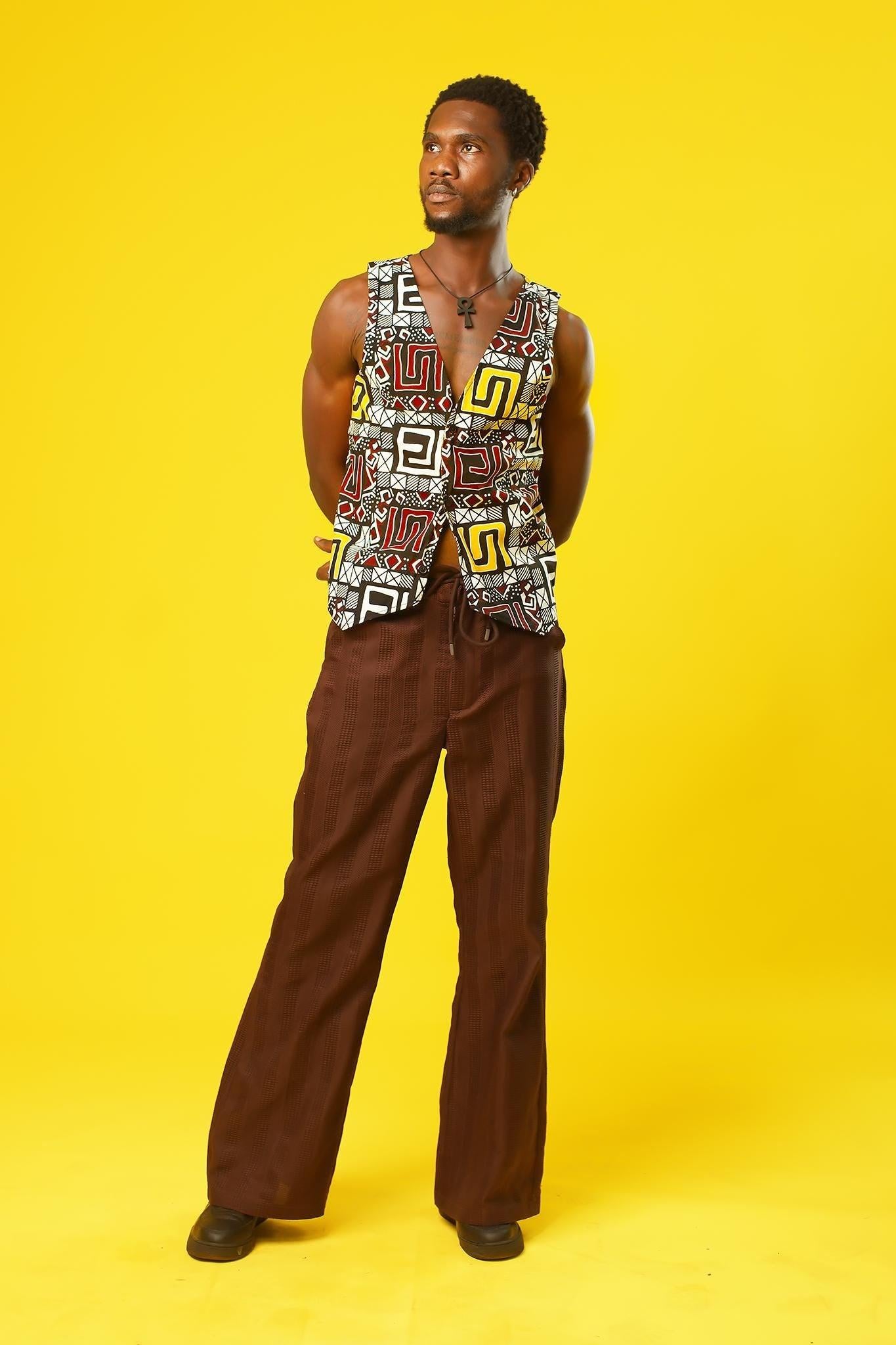 A man models a Reversible Men’s Waistcoat made from bold retro African print fabric, featuring a geometric patchwork of red, yellow, black, and white designs. He pairs the sleeveless vest with relaxed brown drawstring trousers and a black ankh necklace, standing against a bright yellow background. This boho cotton waistcoat is perfect for 70s hippie men’s fashion, festival outfits, or statement Afro-inspired streetwear.