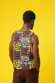 A man models a Reversible Men’s Waistcoat made from bold retro African print fabric, featuring a geometric patchwork of red, yellow, black, and white designs. He pairs the sleeveless vest with relaxed brown drawstring trousers and a black ankh necklace, standing against a bright yellow background. This boho cotton waistcoat is perfect for 70s hippie men’s fashion, festival outfits, or statement Afro-inspired streetwear.