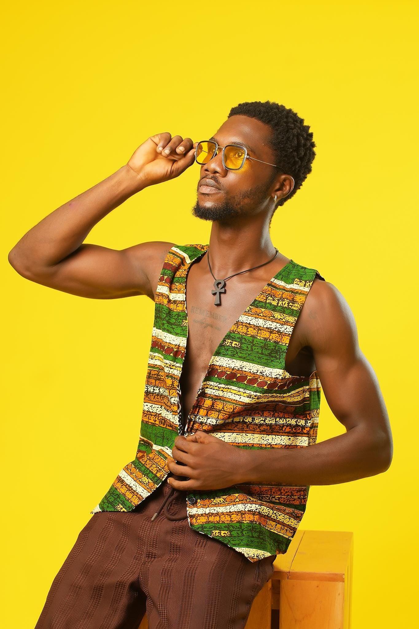 A confident man poses in a Reversible Men’s Waistcoat made from retro African print fabric in rich earthy tones. The handmade cotton vest features a bold horizontal pattern and is styled open over brown trousers. Paired with yellow sunglasses and a black ankh necklace, it’s the perfect festival outfit—ideal for 70s hippie men’s clothing, boho style lovers, or anyone into brown kawaii fashion and Afro-inspired looks.