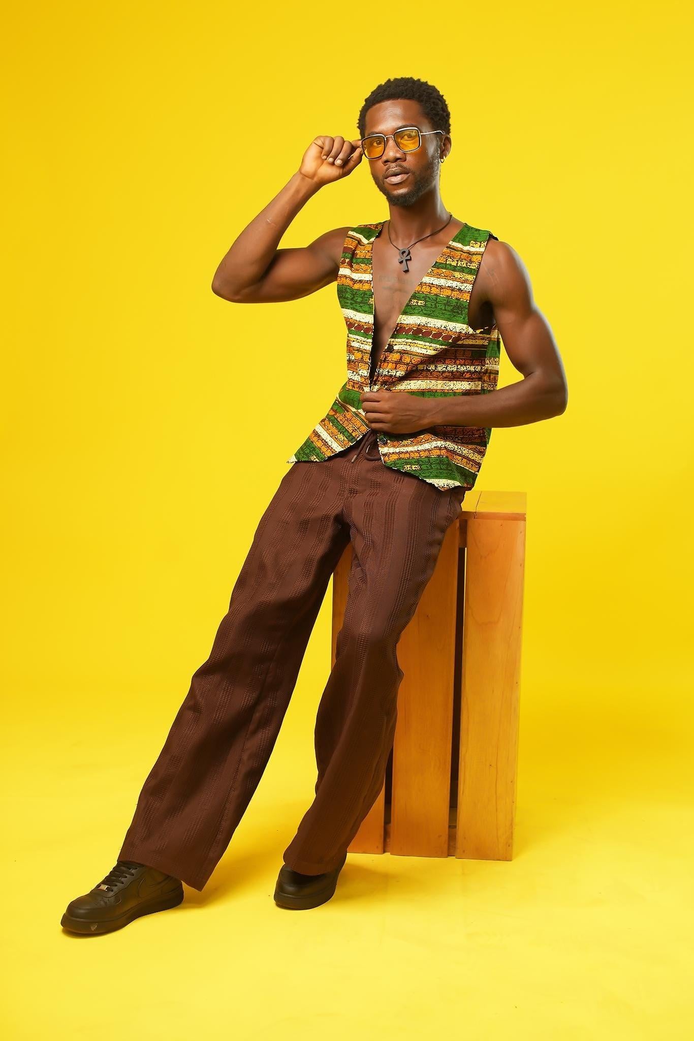 A stylish man poses against a bold yellow background, wearing a sleeveless African wax print vest in earthy green, orange, and brown tones. The handmade vest features traditional strip patterns and is paired with brown textured trousers. He accessorises with yellow-tinted sunglasses and a black ankh necklace, giving a cool, festival-ready vibe. Perfect for modern African fashion, Ankara streetwear, and Afro-inspired style.