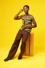 A stylish man poses against a bold yellow background, wearing a sleeveless African wax print vest in earthy green, orange, and brown tones. The handmade vest features traditional strip patterns and is paired with brown textured trousers. He accessorises with yellow-tinted sunglasses and a black ankh necklace, giving a cool, festival-ready vibe. Perfect for modern African fashion, Ankara streetwear, and Afro-inspired style.
