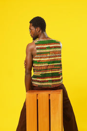 A stylish man poses against a bold yellow background, wearing a sleeveless African wax print vest in earthy green, orange, and brown tones. The handmade vest features traditional strip patterns and is paired with brown textured trousers. He accessorises with yellow-tinted sunglasses and a black ankh necklace, giving a cool, festival-ready vibe. Perfect for modern African fashion, Ankara streetwear, and Afro-inspired style.