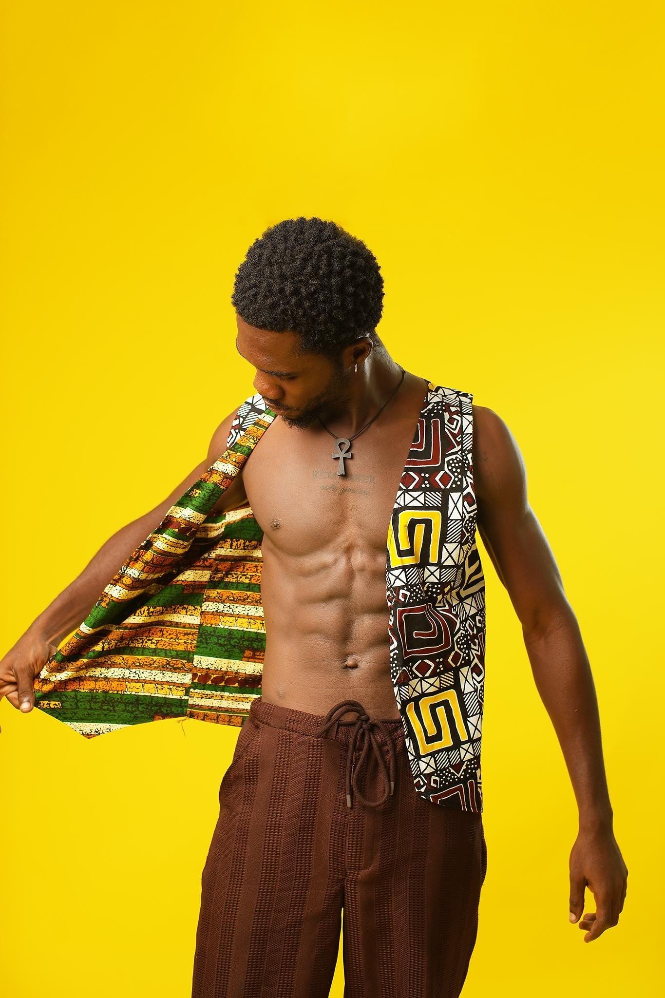 A man models a Reversible Men’s Waistcoat made from bold retro African print fabric, featuring a geometric patchwork of red, yellow, black, and white designs. He pairs the sleeveless vest with relaxed brown drawstring trousers and a black ankh necklace, standing against a bright yellow background. This boho cotton waistcoat is perfect for 70s hippie men’s fashion, festival outfits, or statement Afro-inspired streetwear.