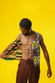A man models a Reversible Men’s Waistcoat made from bold retro African print fabric, featuring a geometric patchwork of red, yellow, black, and white designs. He pairs the sleeveless vest with relaxed brown drawstring trousers and a black ankh necklace, standing against a bright yellow background. This boho cotton waistcoat is perfect for 70s hippie men’s fashion, festival outfits, or statement Afro-inspired streetwear.