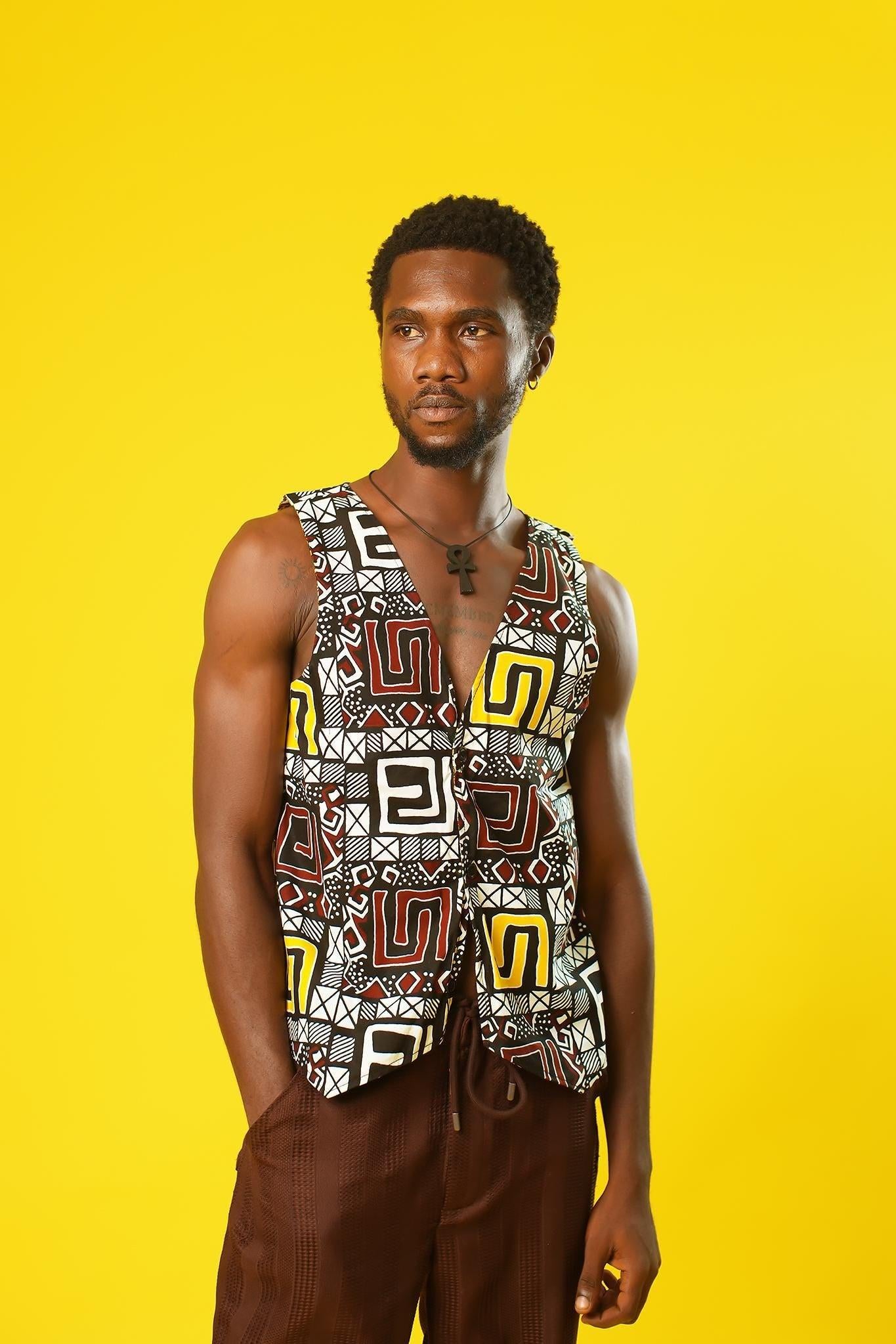 A man models a Reversible Men’s Waistcoat made from bold retro African print fabric, featuring a geometric patchwork of red, yellow, black, and white designs. He pairs the sleeveless vest with relaxed brown drawstring trousers and a black ankh necklace, standing against a bright yellow background. This boho cotton waistcoat is perfect for 70s hippie men’s fashion, festival outfits, or statement Afro-inspired streetwear.
