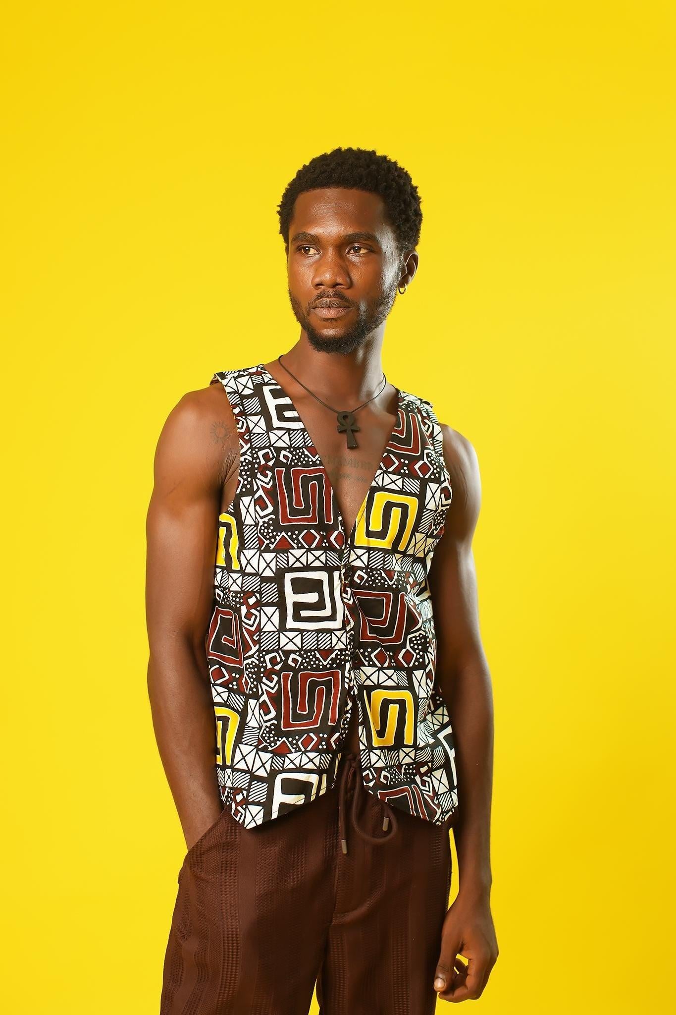 A man models a Reversible Men’s Waistcoat made from bold retro African print fabric, featuring a geometric patchwork of red, yellow, black, and white designs. He pairs the sleeveless vest with relaxed brown drawstring trousers and a black ankh necklace, standing against a bright yellow background. This boho cotton waistcoat is perfect for 70s hippie men’s fashion, festival outfits, or statement Afro-inspired streetwear.