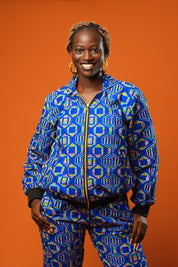 Woman wearing a blue African print tracksuit featuring a zip-up bomber jacket and matching cuffed jogger trousers with yellow, pink, and green geometric batik patterns. The unisex set is styled casually and is ideal for festivals, loungewear, or bold street fashion.