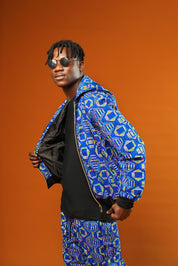 Man wearing a blue African print tracksuit with geometric patterns, featuring a hooded zip-up rave jacket and matching cuffed jogger trousers. The unisex outfit is styled with dark sunglasses and sliders, suitable for disco, festival, or streetwear looks.