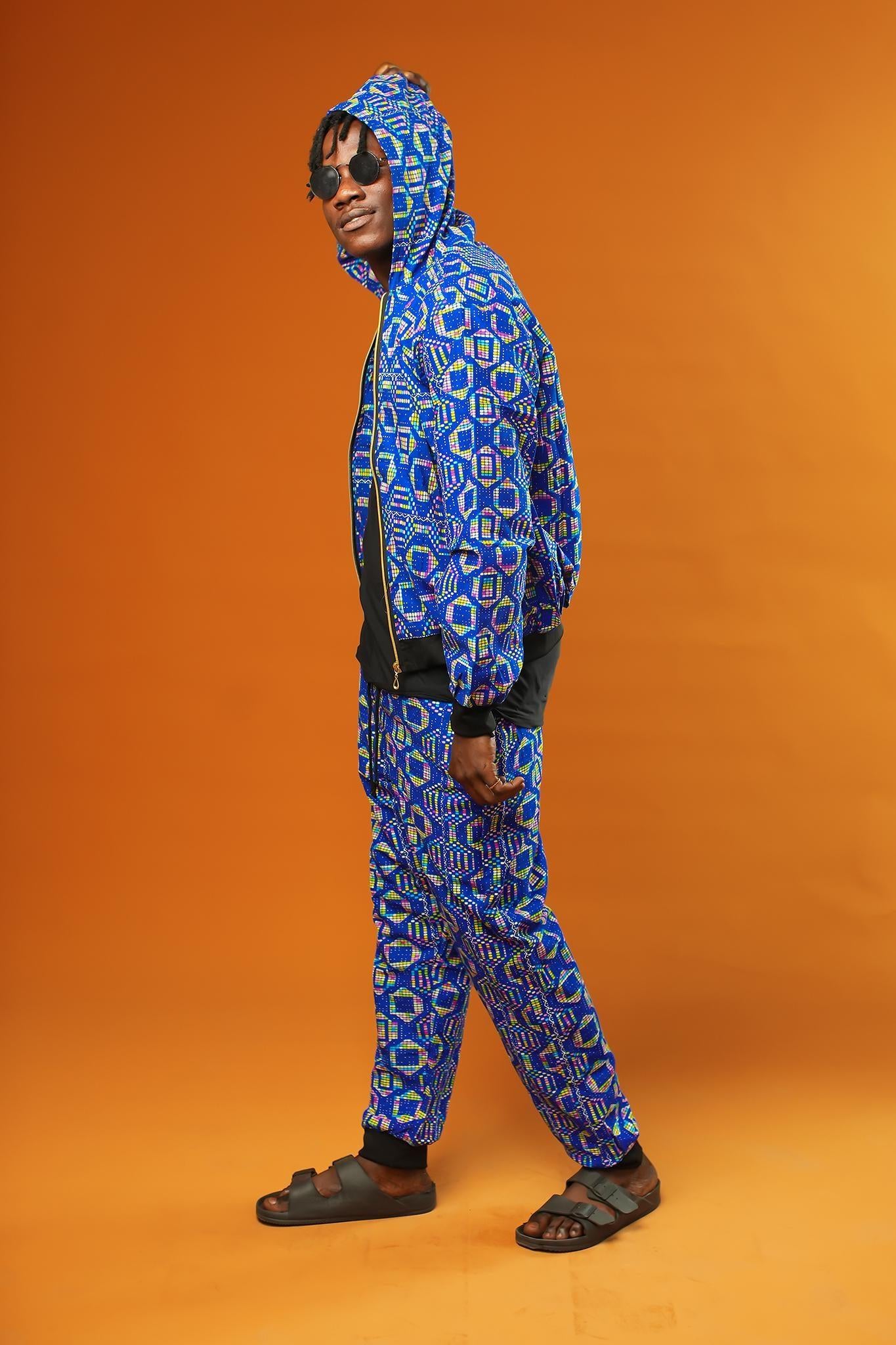 Man wearing a blue African print tracksuit with geometric patterns, featuring a hooded zip-up rave jacket and matching cuffed jogger trousers. The unisex outfit is styled with dark sunglasses and sliders, suitable for disco, festival, or streetwear looks.