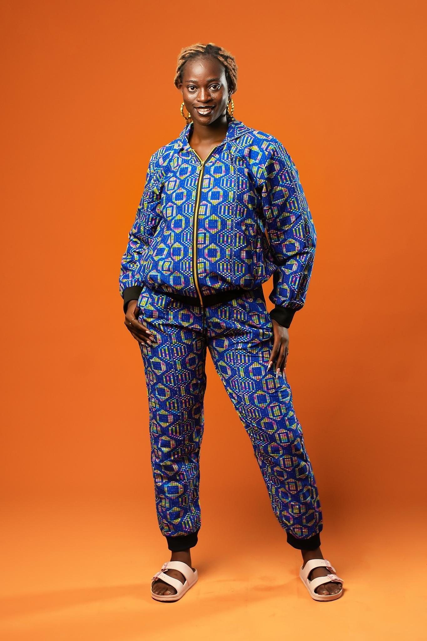Woman wearing a blue African print tracksuit featuring a zip-up bomber jacket and matching cuffed jogger trousers with yellow, pink, and green geometric batik patterns. The unisex set is styled casually and is ideal for festivals, loungewear, or bold street fashion.