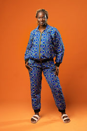 Woman wearing a blue African print tracksuit featuring a zip-up bomber jacket and matching cuffed jogger trousers with yellow, pink, and green geometric batik patterns. The unisex set is styled casually and is ideal for festivals, loungewear, or bold street fashion.