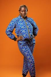 Woman wearing a blue African print tracksuit featuring a zip-up bomber jacket and matching cuffed jogger trousers with yellow, pink, and green geometric batik patterns. The unisex set is styled casually and is ideal for festivals, loungewear, or bold street fashion.