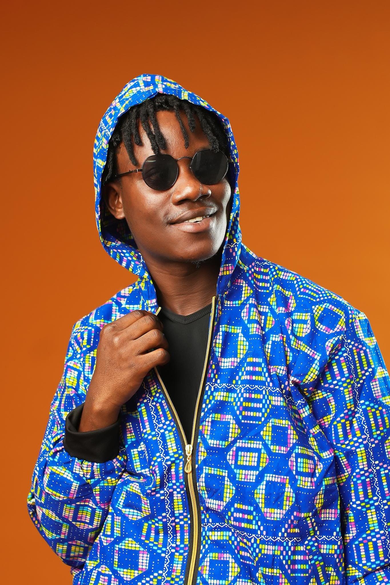 Man wearing a blue African print tracksuit with geometric patterns, featuring a hooded zip-up rave jacket and matching cuffed jogger trousers. The unisex outfit is styled with dark sunglasses and sliders, suitable for disco, festival, or streetwear looks.
