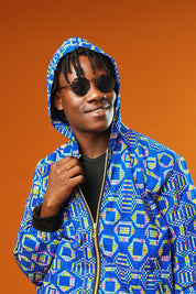 Man wearing a blue African print tracksuit with geometric patterns, featuring a hooded zip-up rave jacket and matching cuffed jogger trousers. The unisex outfit is styled with dark sunglasses and sliders, suitable for disco, festival, or streetwear looks.