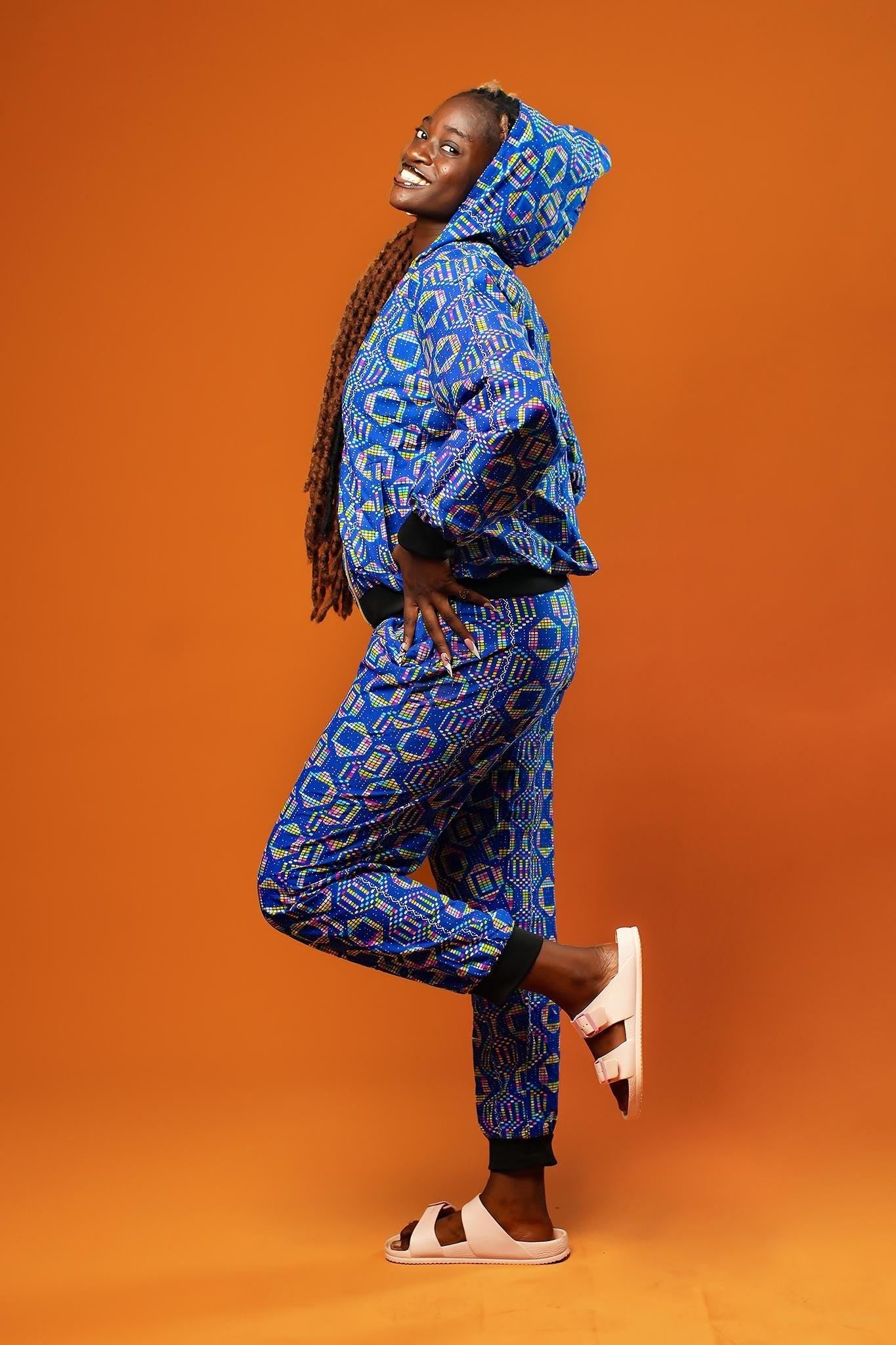 Woman standing against an orange background wearing blue African print jogger trousers with a black drawstring waistband and black ankle cuffs. The trousers feature a colourful geometric pattern in blue, yellow, and red.