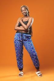 Woman standing against an orange background wearing blue African print jogger trousers with a black drawstring waistband and black ankle cuffs. The trousers feature a colourful geometric pattern in blue, yellow, and red.