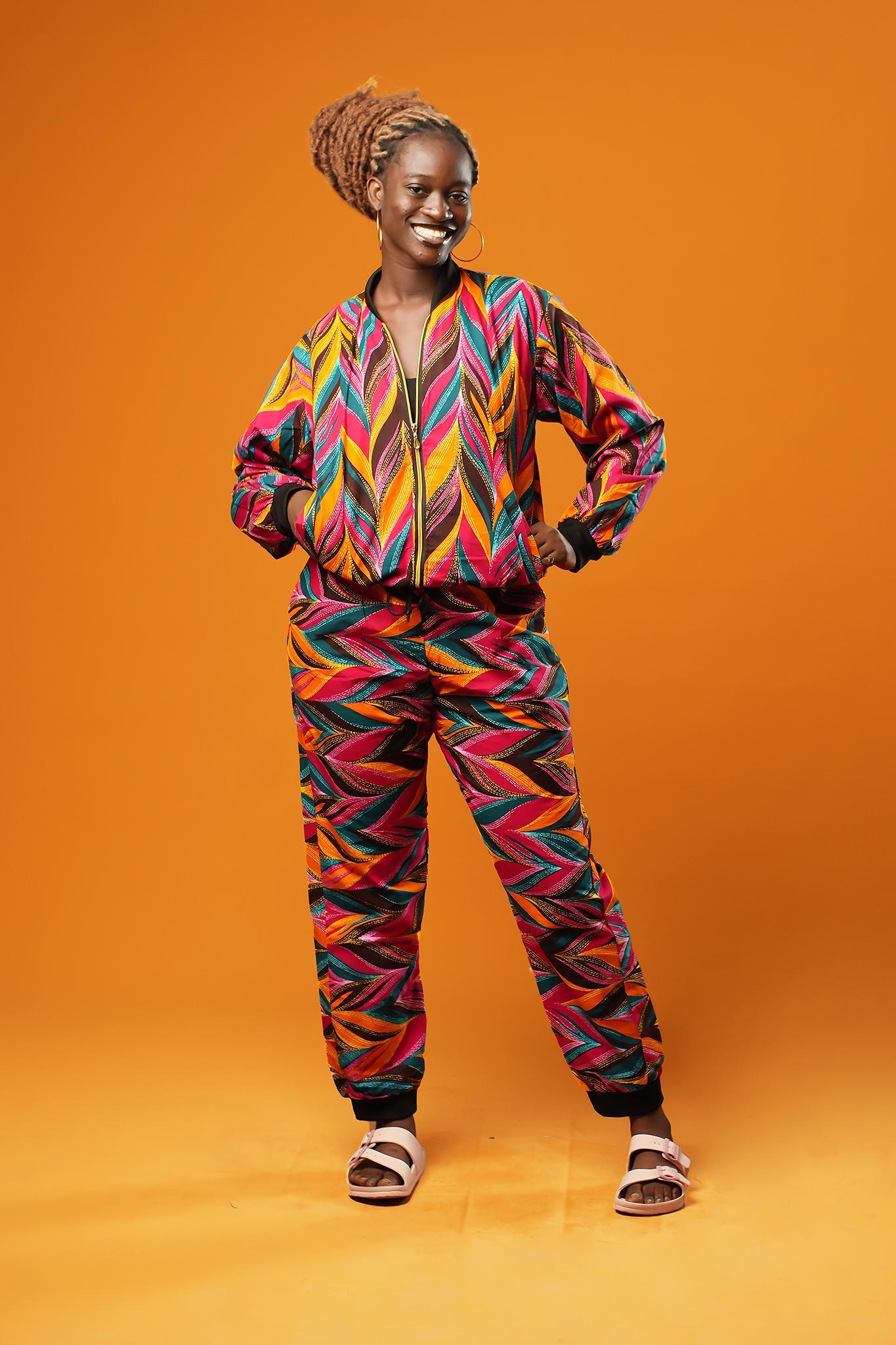 Woman standing against an orange background wearing colourful African Ankara trousers made from bright orange, pink, green, and yellow tribal print fabric. The trousers have a relaxed fit with a black drawstring waistband and black ankle cuffs. Styled as part of a matching tracksuit set. Afrocentric tribal festival rave track pants, lounge pants, by Ashanti Empress.