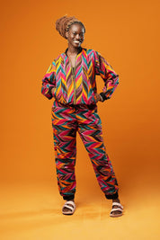 Woman standing against an orange background wearing colourful African Ankara trousers made from bright orange, pink, green, and yellow tribal print fabric. The trousers have a relaxed fit with a black drawstring waistband and black ankle cuffs. Styled as part of a matching tracksuit set. Afrocentric tribal festival rave track pants, lounge pants, by Ashanti Empress.