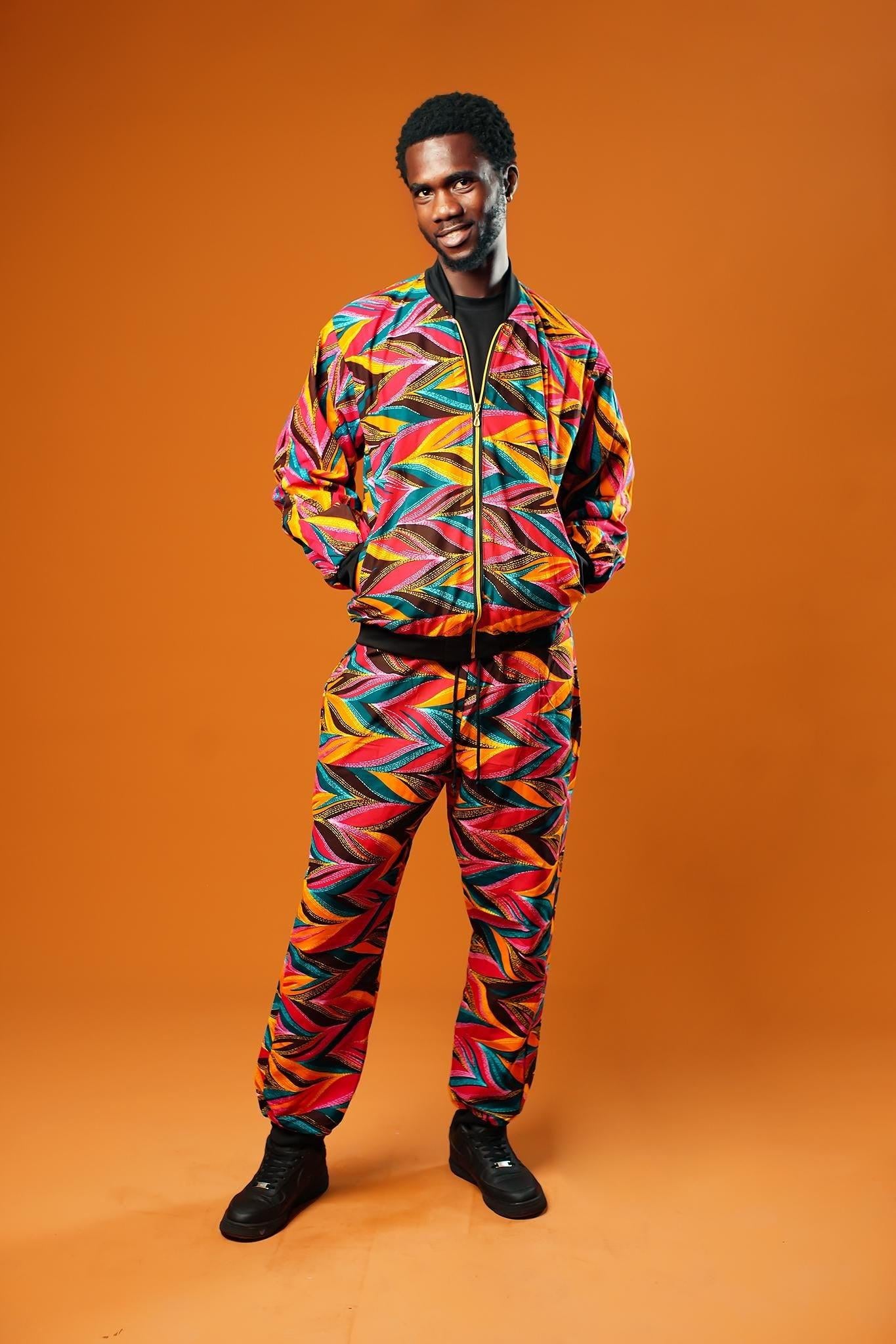 Man standing against an orange background wearing colourful African Ankara trousers made from bright orange, pink, green, and yellow tribal print fabric. The trousers have a relaxed fit with a black drawstring waistband and black ankle cuffs. Styled as part of a matching tracksuit set.