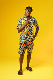 A man stands in front of a yellow background wearing Funky Men’s Cargo Shorts made from multicoloured African print fabric with a butterfly pattern in orange, yellow, blue, and black. The shorts have a relaxed fit and side pockets, paired with a matching short-sleeve shirt. This is a bold Summer Hawaiian Shorts look, ideal for African Print Men’s Clothing, a Hippie Festival Set, or a Carnival Outfit from Ashanti Empress.
