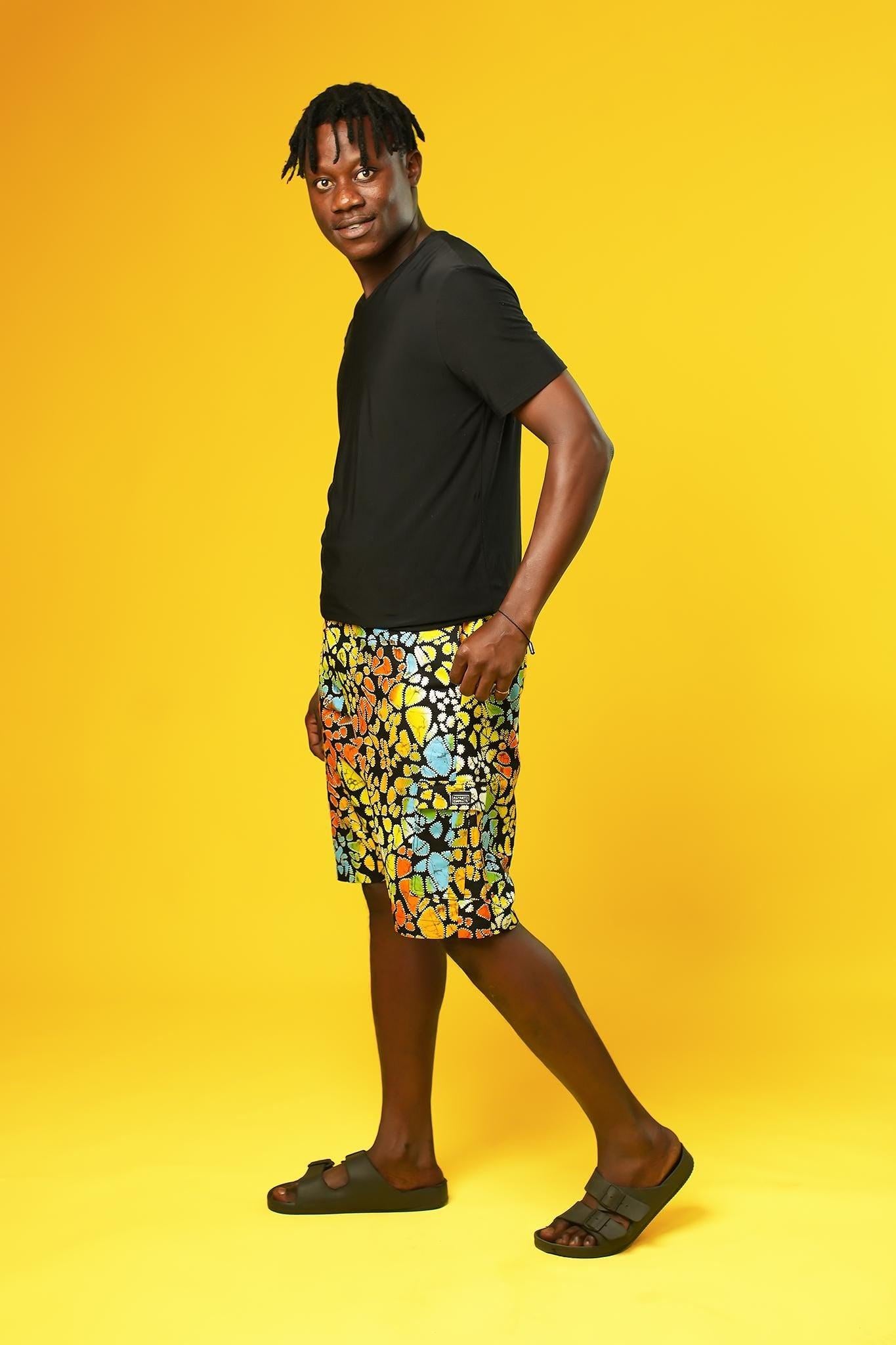 A man stands in front of a yellow background wearing Funky Men’s Cargo Shorts made from multicoloured African print fabric with a butterfly pattern in orange, yellow, blue, and black. The shorts have a relaxed fit and side pockets, paired with a matching short-sleeve shirt. This is a bold Summer Hawaiian Shorts look, ideal for African Print Men’s Clothing, a Hippie Festival Set, or a Carnival Outfit from Ashanti Empress.