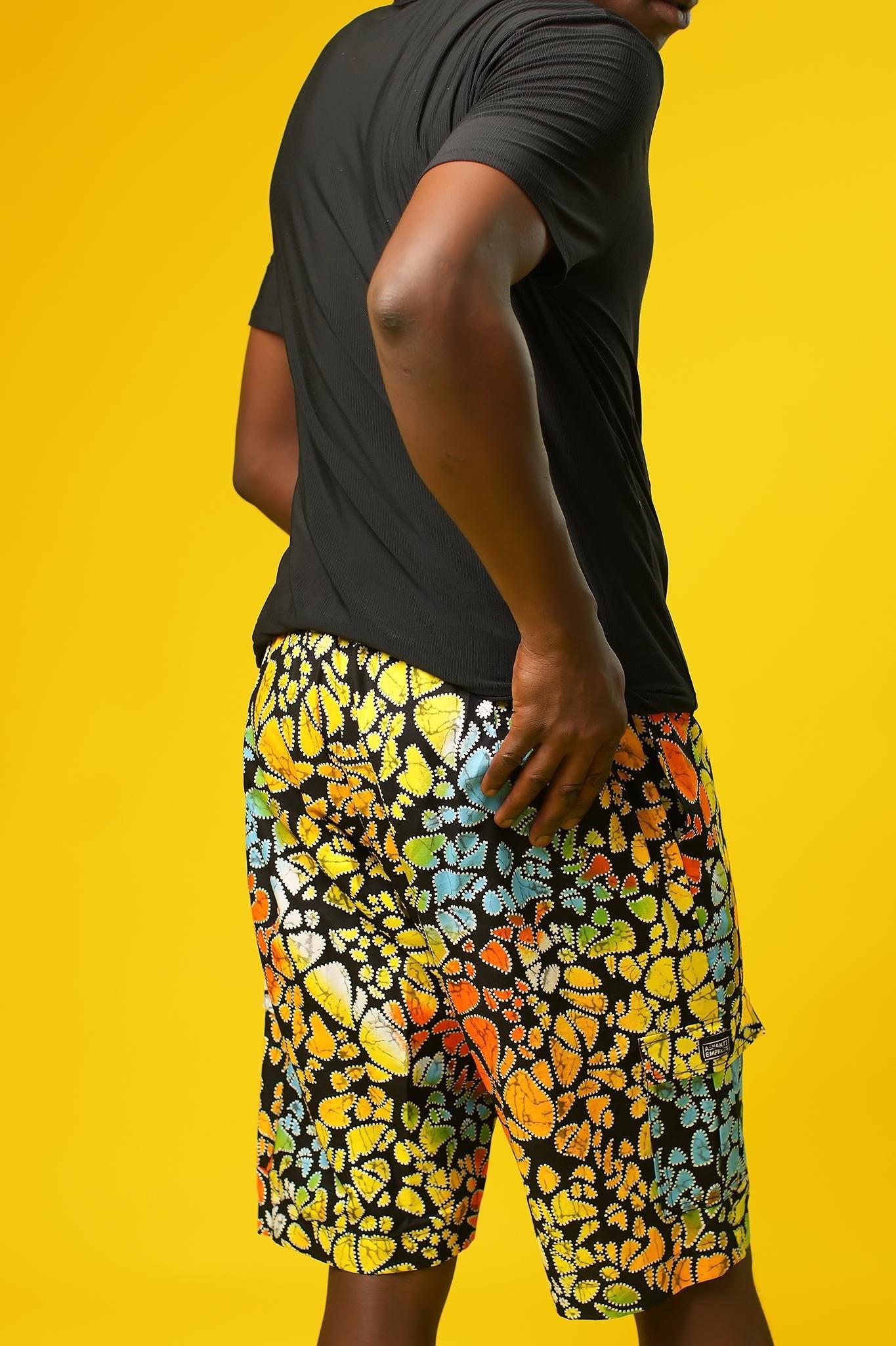 A man stands in front of a yellow background wearing Funky Men’s Cargo Shorts made from multicoloured African print fabric with a butterfly pattern in orange, yellow, blue, and black. The shorts have a relaxed fit and side pockets, paired with a matching short-sleeve shirt. This is a bold Summer Hawaiian Shorts look, ideal for African Print Men’s Clothing, a Hippie Festival Set, or a Carnival Outfit from Ashanti Empress.