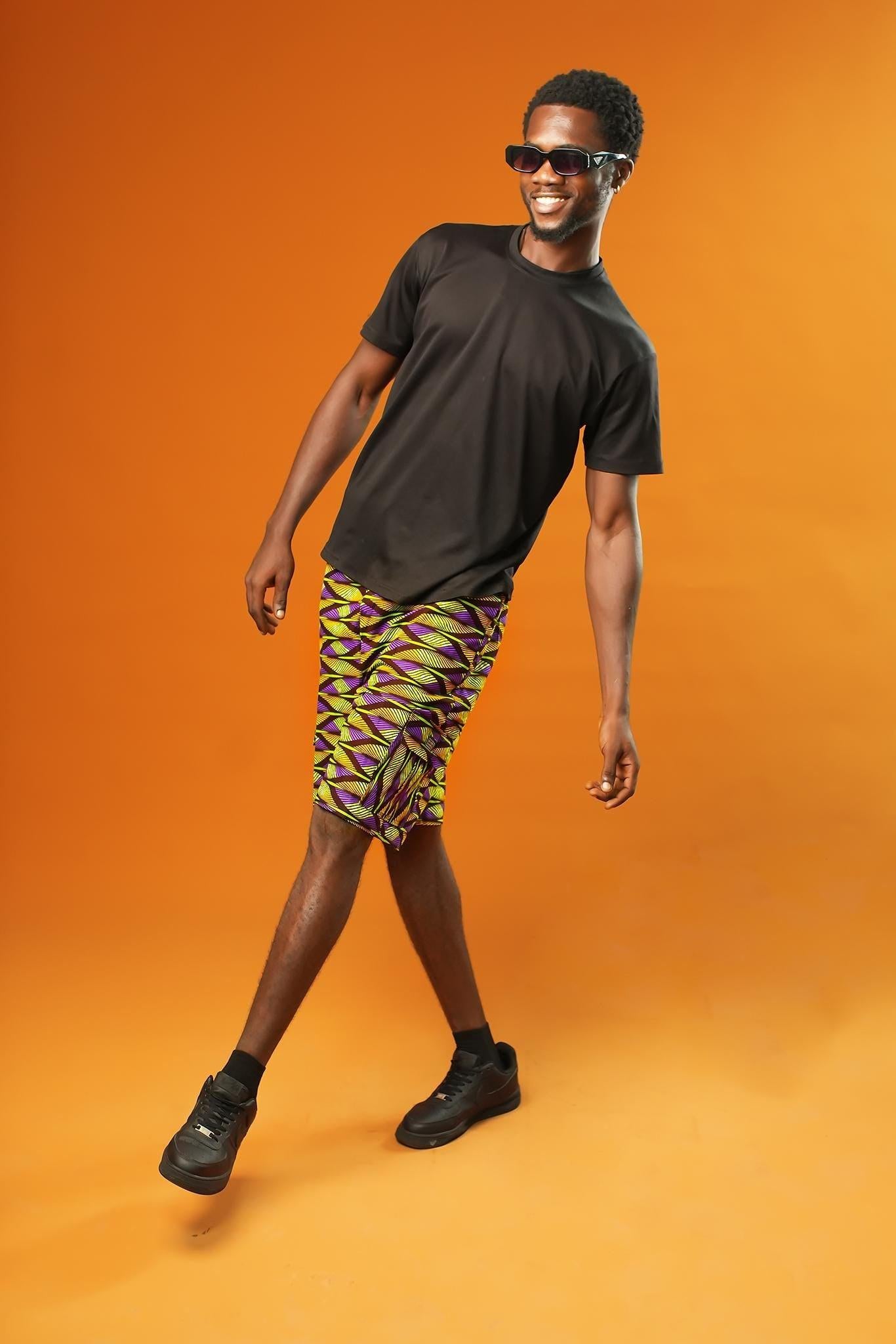 Man wearing Aztec green men’s shorts made from African print cotton fabric with a yellow, black, and purple geometric pattern. The shorts are knee-length with a relaxed fit and side cargo pockets. Paired with a matching shirt, black trainers, and sunglasses. Orange background.