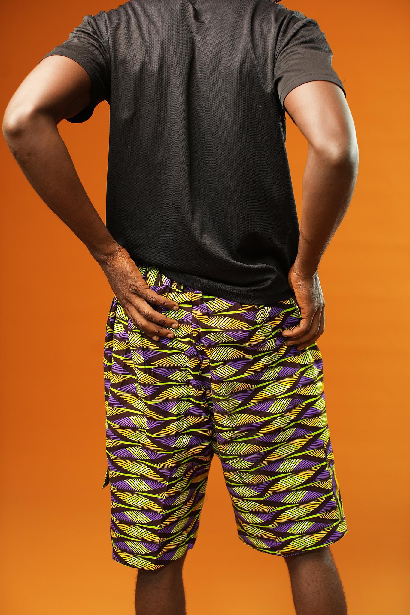 Man wearing Aztec green men’s shorts made from African print cotton fabric with a yellow, black, and purple geometric pattern. The shorts are knee-length with a relaxed fit and side cargo pockets. Paired with a matching shirt, black trainers, and sunglasses. Orange background.